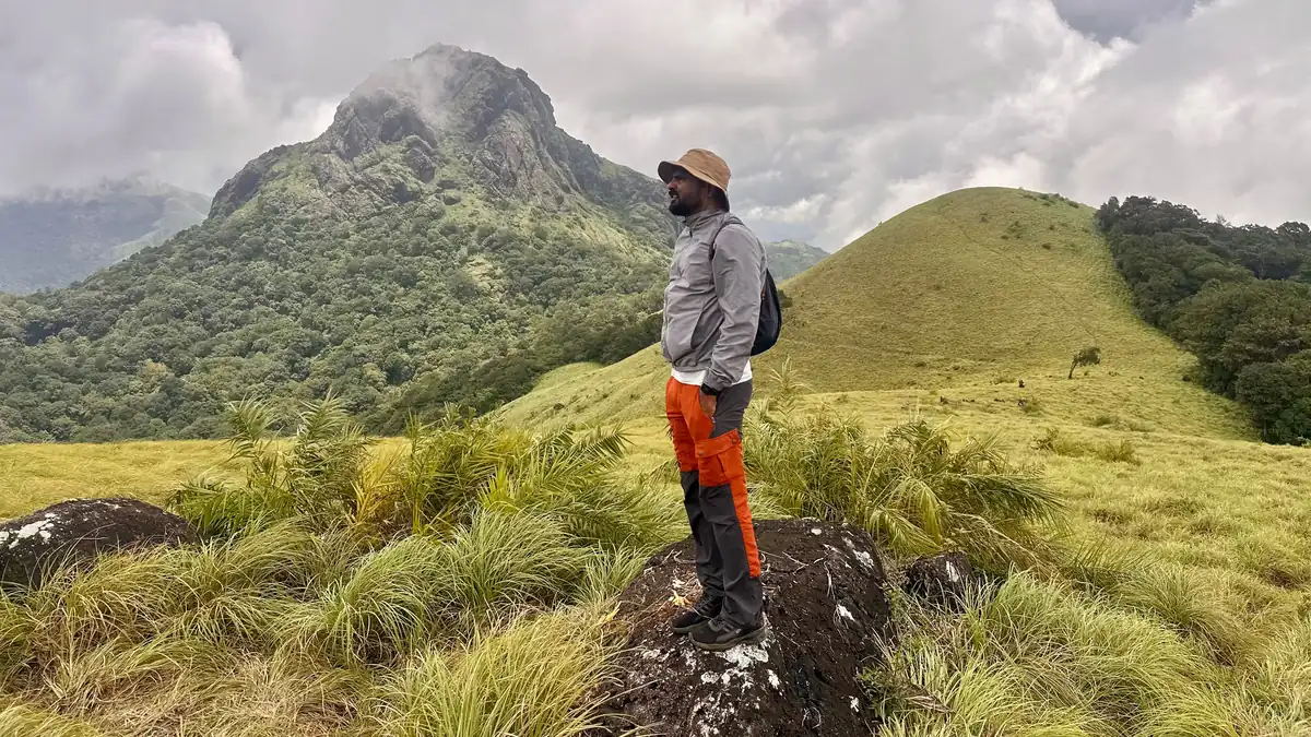 Trekker standing on a rock in the Silent Valley grasslands with a cloud-covered peak behind, Kerala